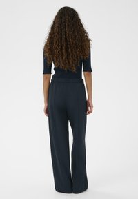 Vrouw met lang, krullend haar, gekleed in een zwarte top met korte mouwen en losse zwarte broek, van achteren afgebeeld tegen een effen witte achtergrond.