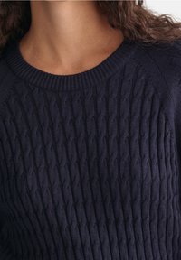 Nahaufnahme einer Person, die einen dunkelblauen Strickpullover mit geripptem Rundhalsausschnitt und lockigem braunem Haar trägt.