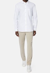 En person står klädd i en vit långärmad skjorta, beige byxor och vita sneakers, mot en neutral bakgrund, vilket visar upp en avslappnad klädsel.