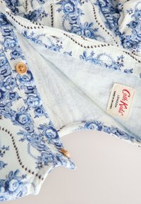 Világoskék alapon kék virágmintás szövet. Egy kis fa gombbal és egy "CarbKids, London" feliratú címkével díszítve.