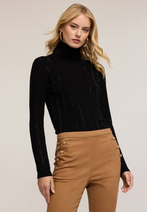 Suéter de cuello alto negro con textura de costillas verticales; acompañado de pantalones de tiro alto color beige que presentan acentos de botones dorados.