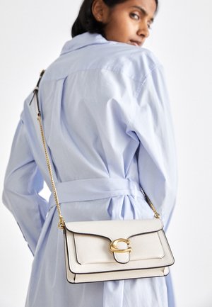Kvinde i lyseblå frakke med bælte, der bærer en hvid læder crossbody taske med guldlås og kædestrop over skulderen.