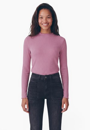 Femme souriante aux longs cheveux foncés portant un haut rose côtelé à manches longues, ajusté, et un jean noir taille haute sur fond blanc.