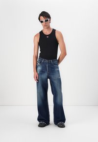 Svart linne med en liten röd logotyp, tillsammans med vida blå denimjeans med urtvättade områden och en fransad fåll. Vita solglasögon fulländar looken.