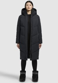 Sort polstret lang frakke med hætte, der har et quiltet design og lynlås. Båret med sorte leggings og blanke sorte støvler.