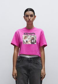 Roze cropped t-shirt met korte mouwen, voorzien van een geprinte afbeelding en het woord "Clueless" in het zwart. Gecombineerd met donkergrijze high-waisted jeans.
