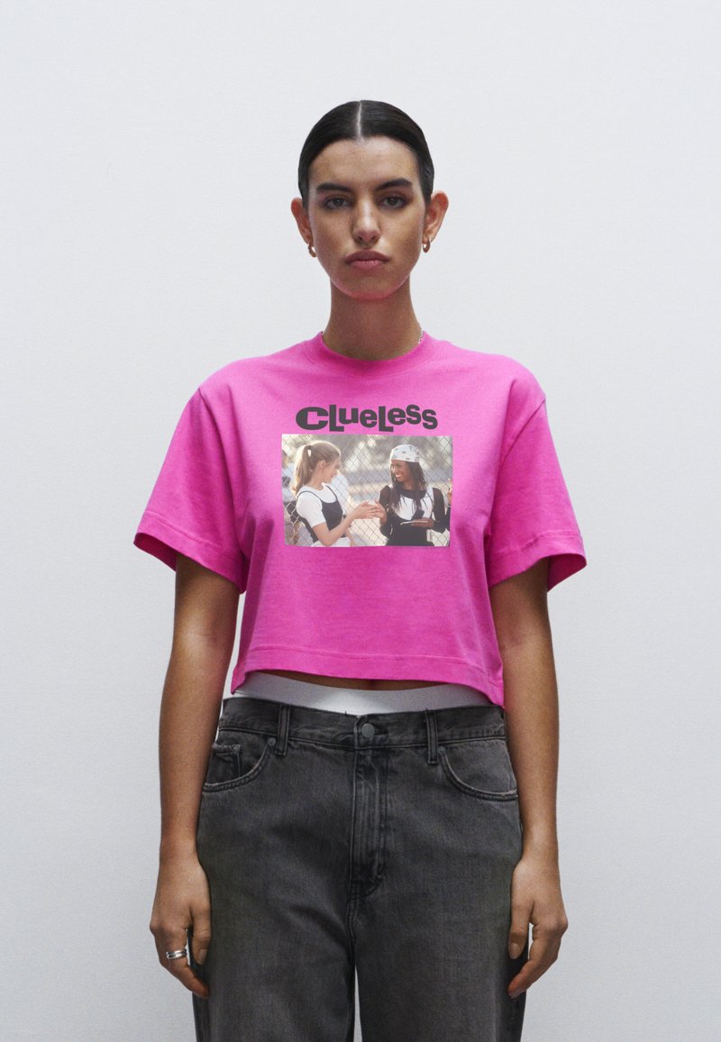 Roze cropped t-shirt met korte mouwen, voorzien van een geprinte afbeelding en het woord "Clueless" in het zwart. Gecombineerd met donkergrijze high-waisted jeans.