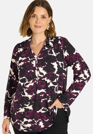 Frau mit kurzen braunen Haaren trägt eine langärmelige, lila-schwarze Bluse mit Blumenmuster und schwarze Hose, Hand in der Tasche, neutraler Hintergrund.