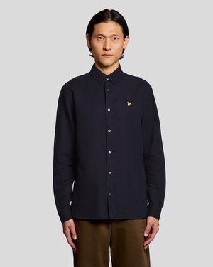 Bărbat purtând o cămașă neagră cu nasturi și un logo galben cu o pasăre pe piept, stând în fața unui fundal simplu de culoare gri deschis.