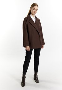 Bruine oversized jas met een insnijding in de kraag, enkele knoopsluiting en brede mouwen, gecombineerd met zwarte broek en enkellaarsjes.