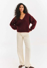 Bordowy sweter z dzianiny z głębokim dekoltem w serek i szerokimi rękawami, zestawiony z kremowymi rozszerzanymi spodniami i brązowymi trampkami.