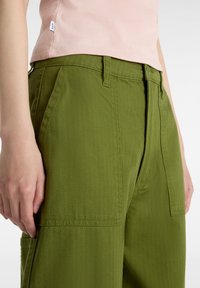 Olivgröna bomullbyxor med texturerad yta, som har framfickor, dragkedja och en liten etikett vid midjan.