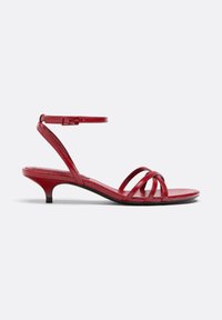 Sandale rouge en cuir avec un petit talon kitten bas, fines lanières croisées et une bride de cheville réglable avec boucle sur fond blanc.