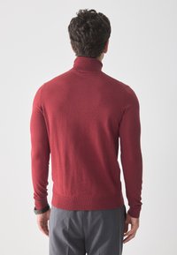 Pull à col roulé rouge en maille douce, avec des manches longues ajustées et un ourlet côtelé. Porté avec un pantalon gris.