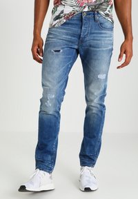 Modré džíny z denimu s úzkým střihem, s opotřebením a vyblednutím na přední straně, doplněné bílými teniskami.