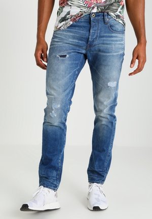 Vaqueros de mezclilla azul con un corte ajustado, con desgastes y decoloraciones en la parte frontal, combinados con zapatillas blancas.