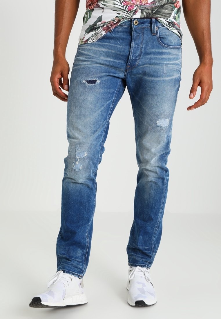 Modré džíny z denimu s úzkým střihem, s opotřebením a vyblednutím na přední straně, doplněné bílými teniskami.