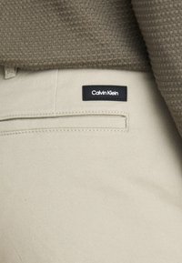 Bež hlače s sleek dizajnom, z zadnjim žepom in črno etiketko Calvin Klein. Teksturovani olivno zeleni pulover je delno viden zgoraj.