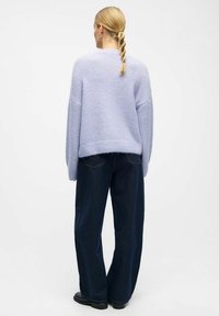 Jasnofioletowy, oversize sweter o miękkiej fakturze, z prążkowanym obszyciem i opuszczonymi ramionami, zestawiony z ciemnymi jeansami o szerokich nogawkach i czarnymi butami.