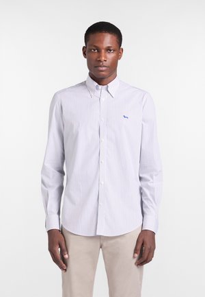 Joven con camisa blanca de botones con finas rayas verticales y pantalones beige, de pie frente a un fondo blanco liso.