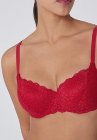 Červená krajková podprsenka s květinovými vzory, s kosticemi a nastavitelnými ramínky. Zubatý výstřih dodává designu detail.