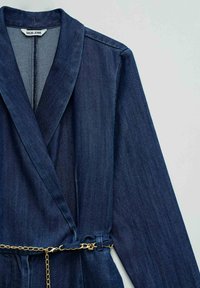 Giacca di denim blu scuro con maniche lunghe, collo a scialle e una sottile cintura a catena dorata in vita.