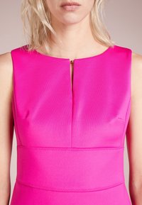 Kvinna i en ärmlös klännings i starkt rosa med en liten öppning fram och bred midjebanddetalj, visad från hals till midja.