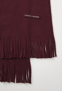 Sciarpa lavorata a maglia bordeaux con bordi frangiati e un'etichetta con il marchio "TOMMY HILFIGER." Texture morbida con una finitura liscia.