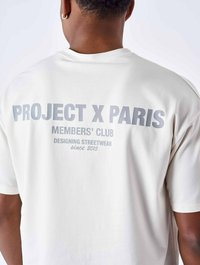 Witte T-shirt met korte mouwen met grijze bedrukte tekst op de achterkant: "PROJECT X PARIS," "MEMBERS' CLUB," en "DESIGNING STREETWEAR since 2015."