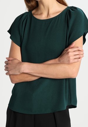 Femme portant un chemisier à manches courtes vert foncé avec des détails froncés aux épaules, les bras croisés, sur un fond clair uni.