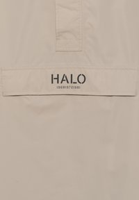 Beżowa odzież wierzchnia z gładkiej, lekkiej tkaniny. Posiada naszywkę z logo "HALO" oraz czarne cyfry 196819721981.