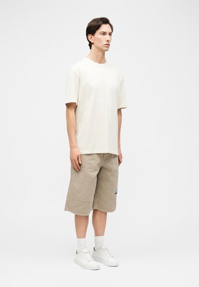 Jeune homme debout, portant un t-shirt à manches courtes uni de couleur blanc cassé, un short beige jusqu'aux genoux avec des poches plaquées, des chaussettes blanches et des baskets blanches.