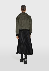 Olivovo zelená imitácia kožušiny bomber bundy s čiernymi rebrovanými manžetami, skombinovaná s čiernou plisovanou midi sukňou. Modelka má obuté čierne členkové čižmy.