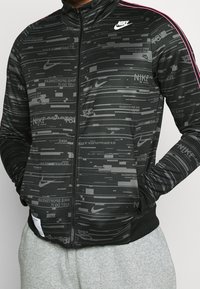Svart zip-up jacka med en texturerad design som har upprepade Nike-loggor i olika nyanser. Rosa accenter på axlarna.