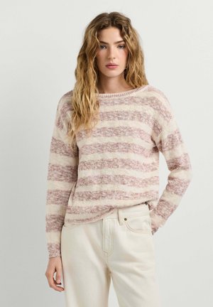 Jeune femme aux longs cheveux ondulés portant un pull rayé crème et rose poudré ainsi qu’un pantalon écru sur un fond uni.