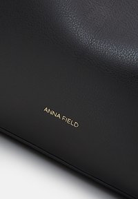 Svart läderhandväska med en texturerad yta. Varumärket "ANNA FIELD" är präglat i guld på den nedre delen.