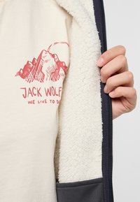 Beige Fleece-Jacke mit blauer Reißverschluss, die ein rotes Bergmotiv und den Text "JACK WOLFKIN" auf hellem Stoff zeigt.
