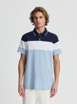 Giovane uomo con capelli ricci che indossa una polo a maniche corte con righe orizzontali blu navy, bianche e azzurro chiaro, in piedi davanti a uno sfondo uniforme.