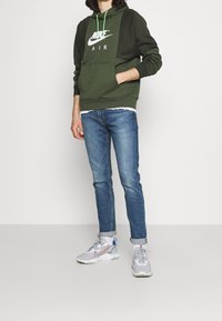 Felpa con cappuccio Nike verde oliva con logo bianco, jeans blu e sneakers Nike grigie. La felpa ha una tasca frontale e un cordino regolabile.