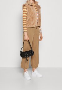 Beige, ärmlös, pälsväst över en randig långärmad skjorta, ihop med tanbyxor och vita sneakers, håller en liten svart handväska.