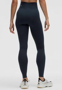 Leggings taille haute, ajustés, en tissu bleu marine foncé, avec une texture lisse, un logo discret et associés à des chaussures de sport blanches.