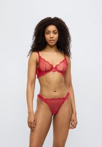 Σετ κόκκινης δαντελένιας εσωρούχων που περιλαμβάνει σουτιέν με κυματιστό σχέδιο και τανγκάκι, με πουά μοτίβα και διάφανη υφή. Λουριά και διακοσμητικές βολάν.