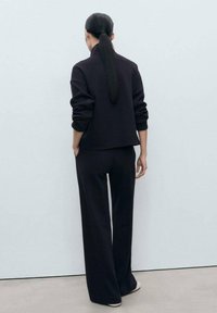 Sweatshirt en maille noire et pantalon large vus de derrière, présentant une coupe décontractée et des manches longues, sur un fond pâle.