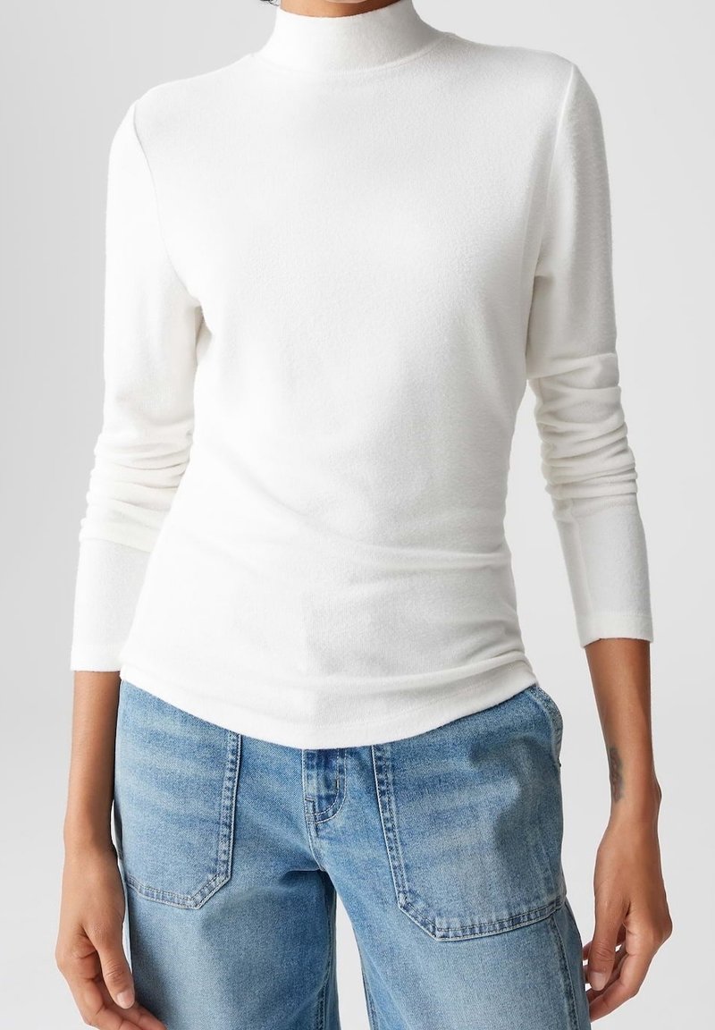 Haut blanc ajusté à col roulé manches longues associé à un jean bleu clair taille haute, présenté sur une personne du cou aux hanches.