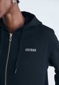 Close-up van een persoon in een zwarte rits-hoodie met het "GUESS" logo op de borst en hangende koordjes.