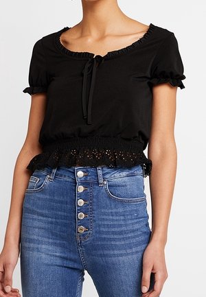 Musta lyhythihainen paita, jossa on röyhelöreunainen pääntie ja smokkivyötärö, solmimisnauhalla ja kirjailtulla reunaosalla. Yhdistetty korkeavyötäröisiin sinisiin farkkuihin.