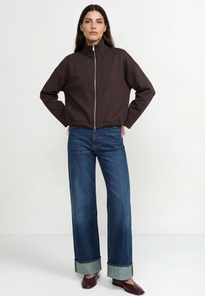 Braune Reißverschlussjacke mit hohem Kragen und elastischem Bund; kombiniert mit weit geschnittenen blauen Jeans, die unten umgeschlagen sind, und dunklen Schuhen.