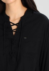 Black shirt featuring a lace-up front, long sleeves, and a chest pocket. Made of a soft material with a relaxed fit. Label visible.