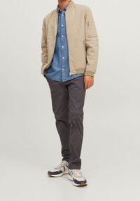 Beige bomberjacka med dragkedjefickor, kombinerad med en ljusblå denimskjorta, grå byxor och flerfärgade sneakers. Avslappnad stil.