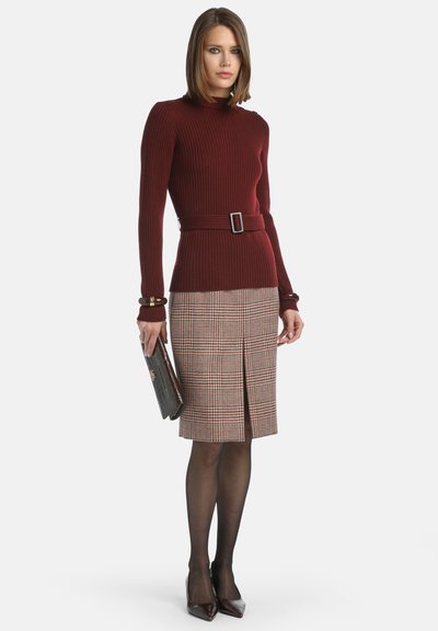 Pull en bourgogne à côtes avec une taille ajustée et une ceinture ; jupe plaid portée jusqu'aux genoux dans des tons de marron et de rouge ; tenant une pochette noire, portant des escarpins pointus.